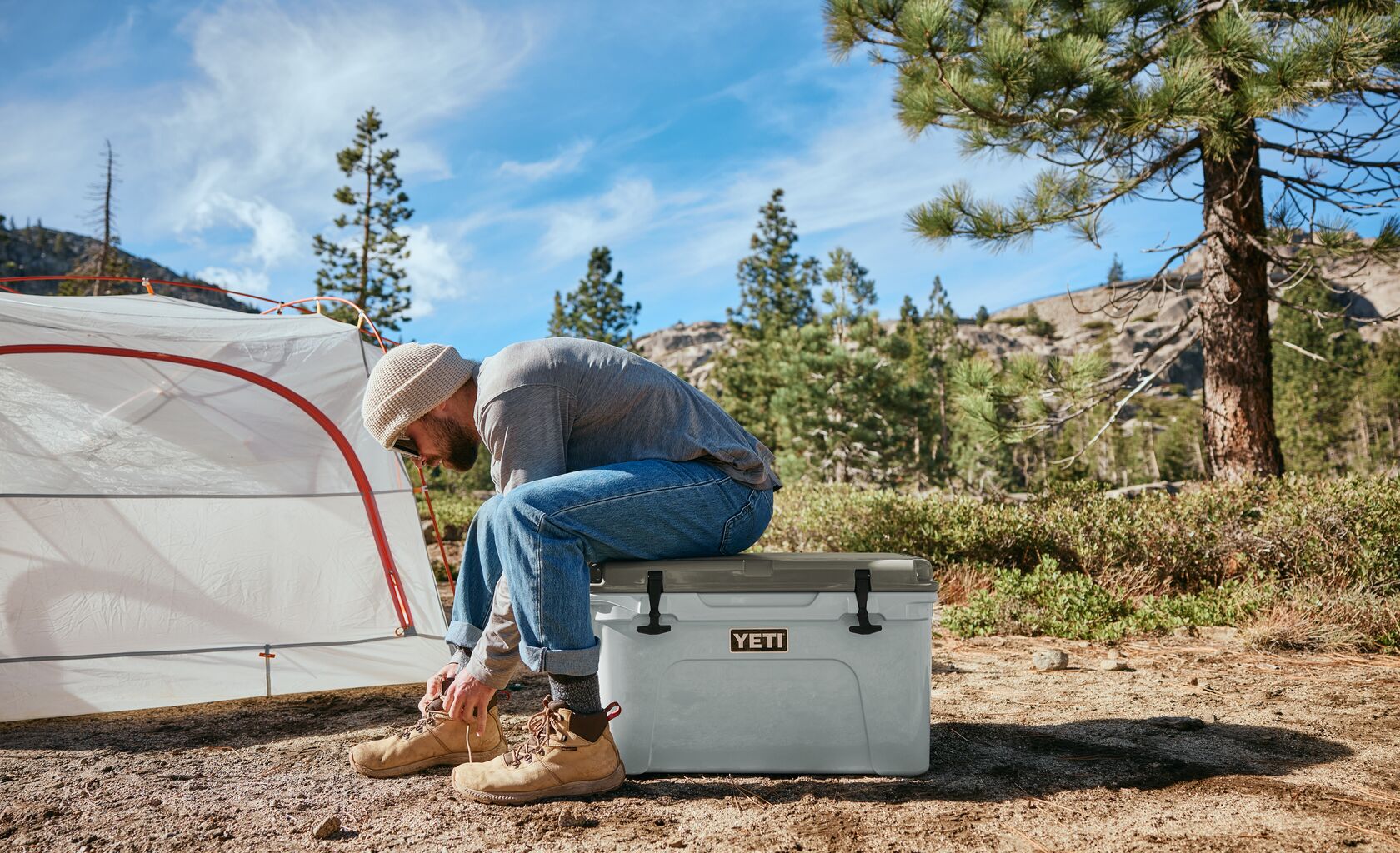Person sitting on a YETI cooler outdoors with a tent and trees in the background